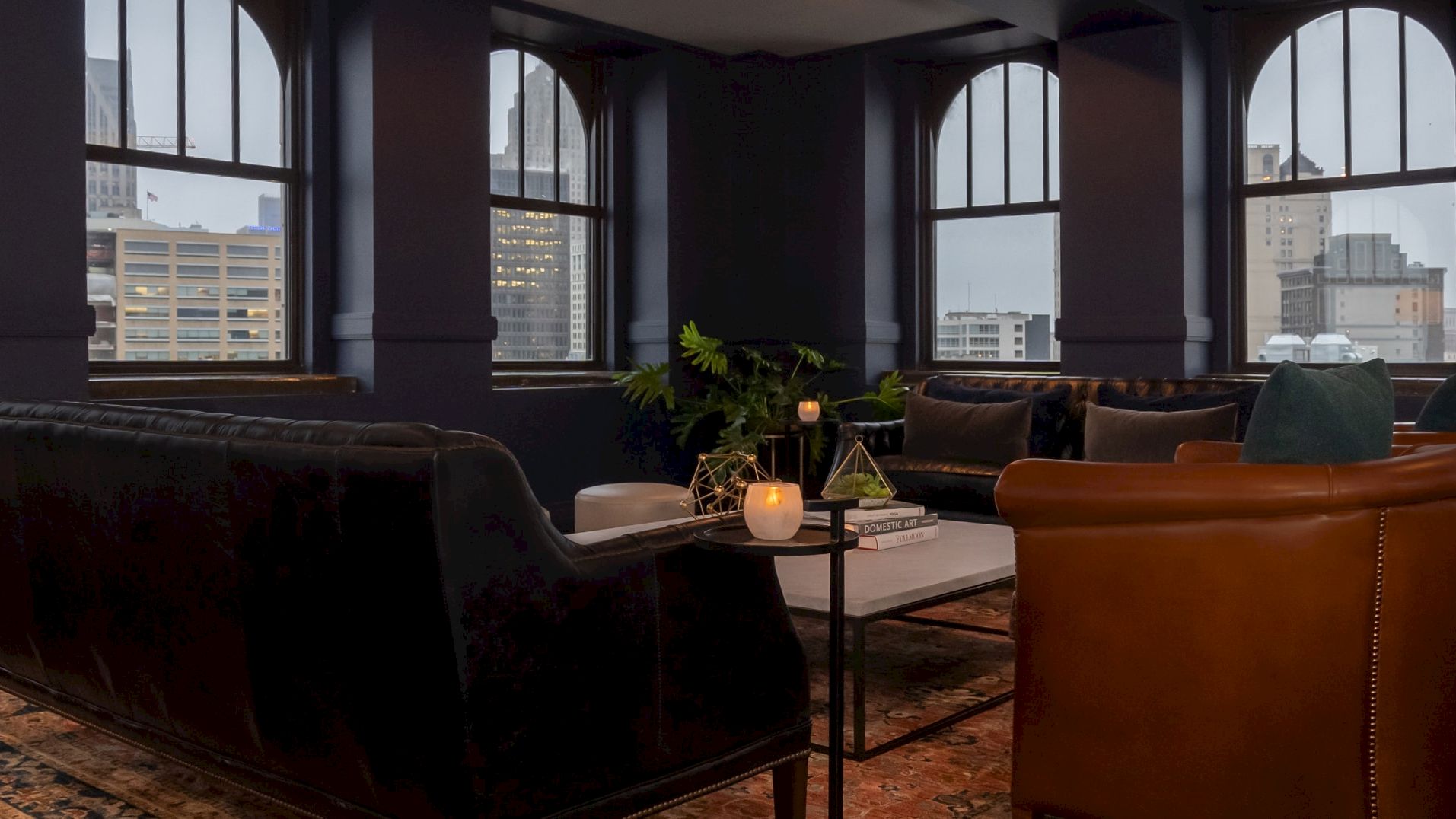 A modern living room with large windows, leather sofas, a geometric coffee table, candles, books, and a plant, offering a city view.