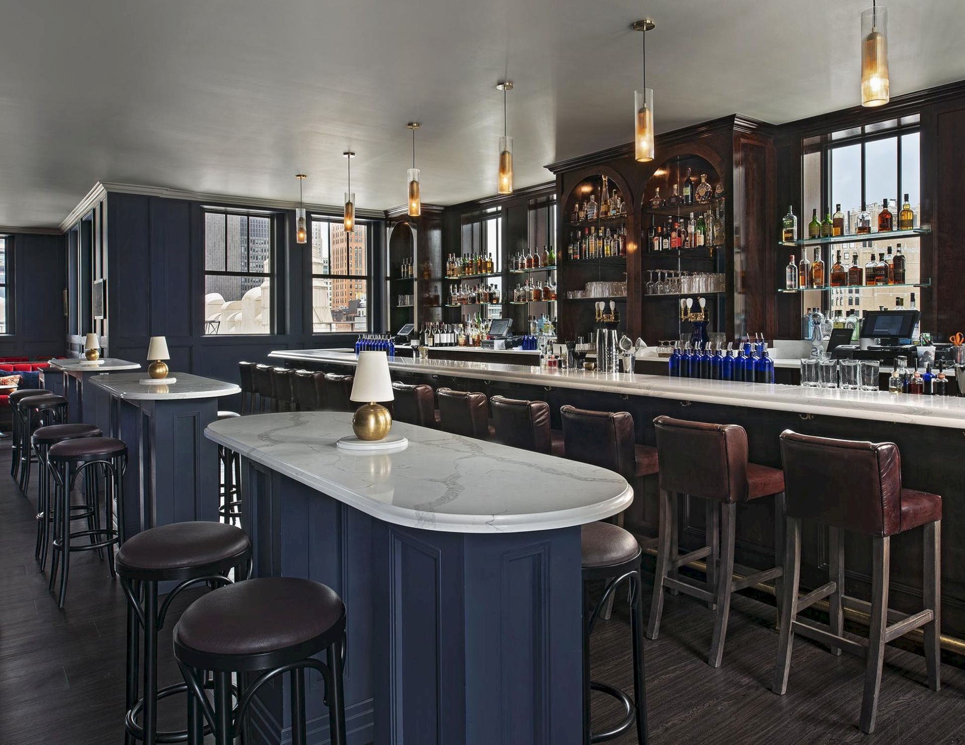 A stylish bar with a long curved white-marble counter, navy paneling, high stools, pendant lights, and shelves stocked with bottles behind the bar, all in a dim, cozy setting.