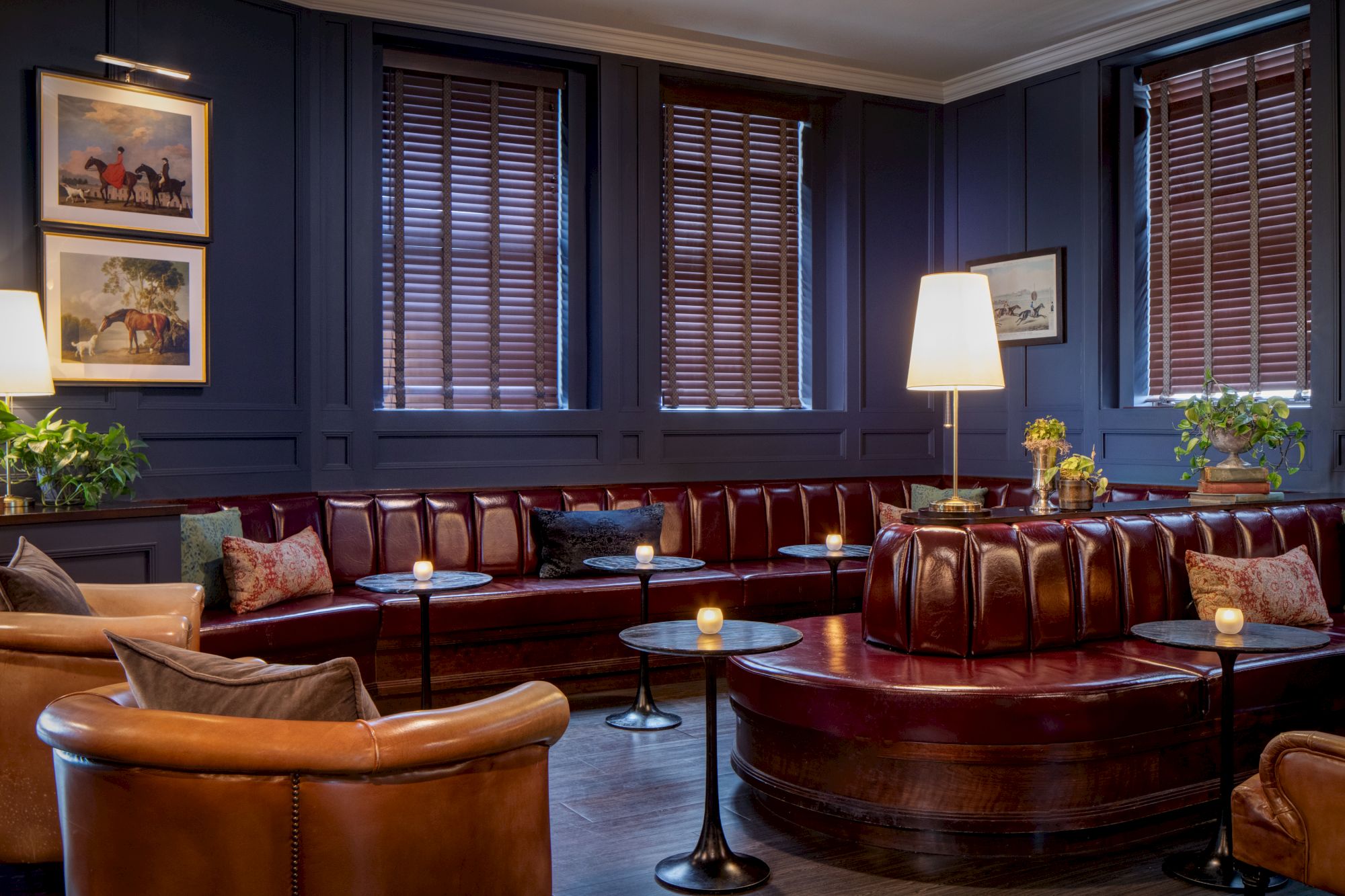 An upscale lounge with brown leather seating, round tables, lamps, framed equestrian art, and large window blinds.