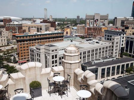 A rooftop terrace with tables and chairs overlooks an urban cityscape featuring various buildings and a distant stadium.