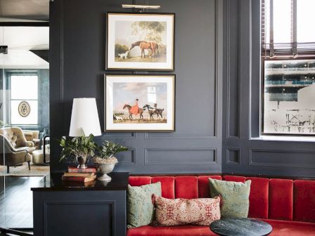 A stylish room with dark walls, red velvet seating, framed horse paintings, a plant, and a lamp near a window with blinds.