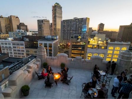 A rooftop gathering at sunset with people seated near a fire pit, surrounded by city buildings reflecting evening lights.