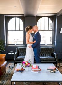 A bride and groom kiss in a modern lounge, surrounded by sofas, arched windows, and wedding gifts on a coffee table.