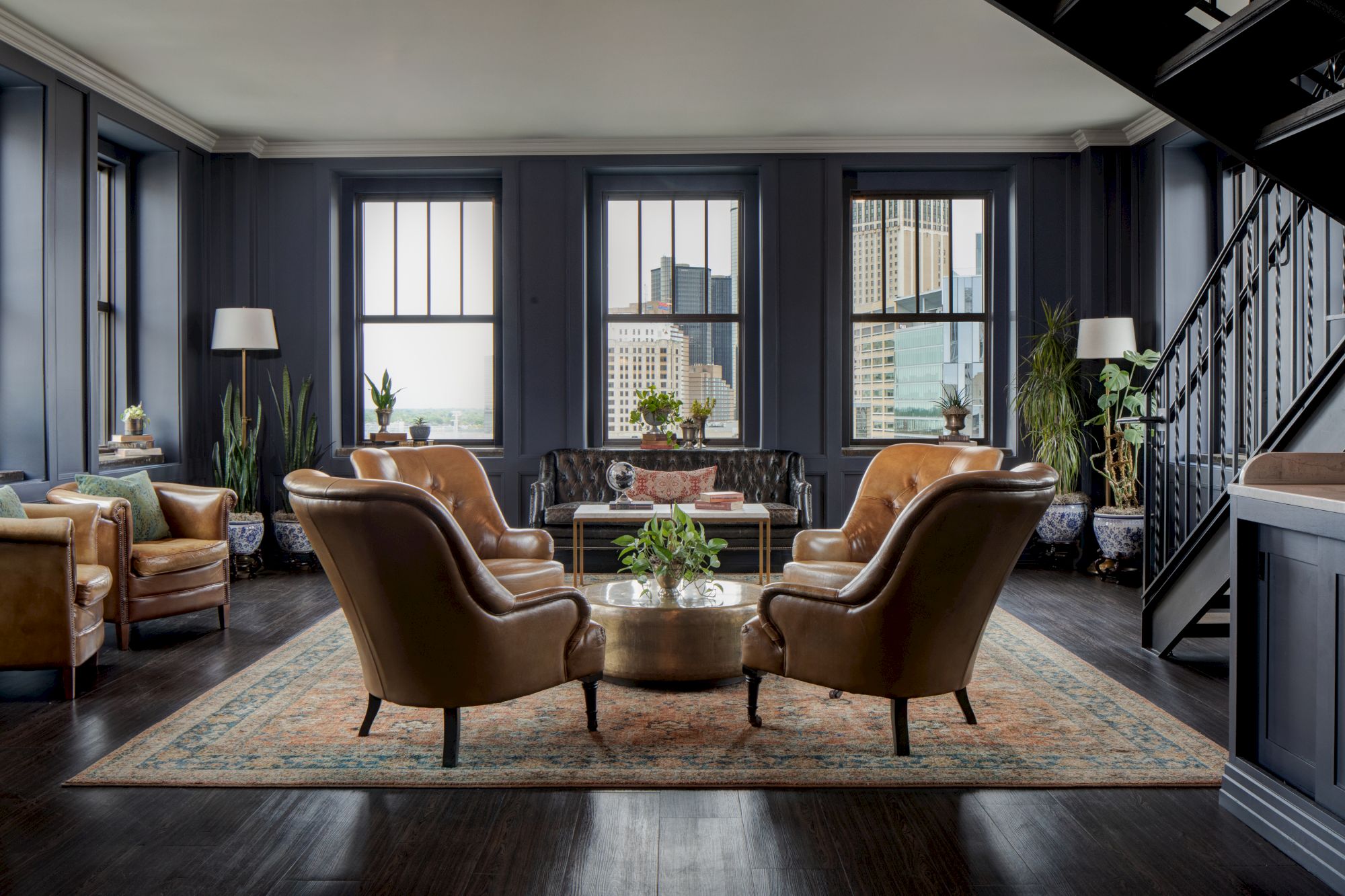 Elegant living room with leather chairs, a central coffee table, indoor plants, and a city view through large windows.