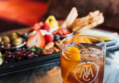 A glass of amber cocktail sits on a table beside a platter of grilled meats and colorful fruit/olives.