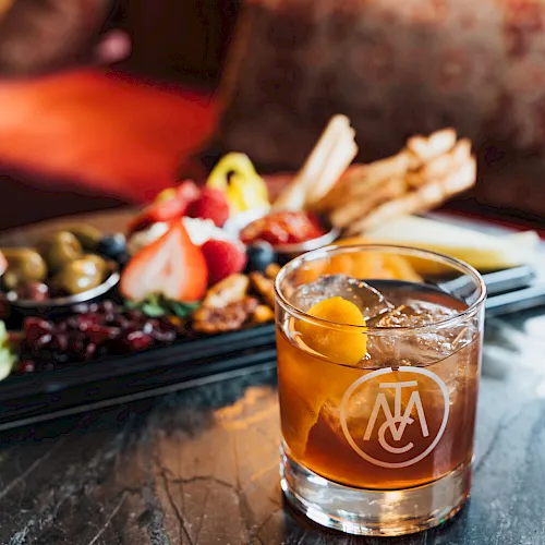 A glass of amber cocktail sits on a table beside a platter of grilled meats and colorful fruit/olives.
