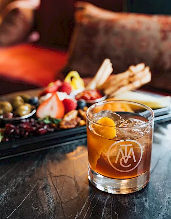 Amber cocktail with ice beside a snack board of fruits, olives, and meats on a rustic table.